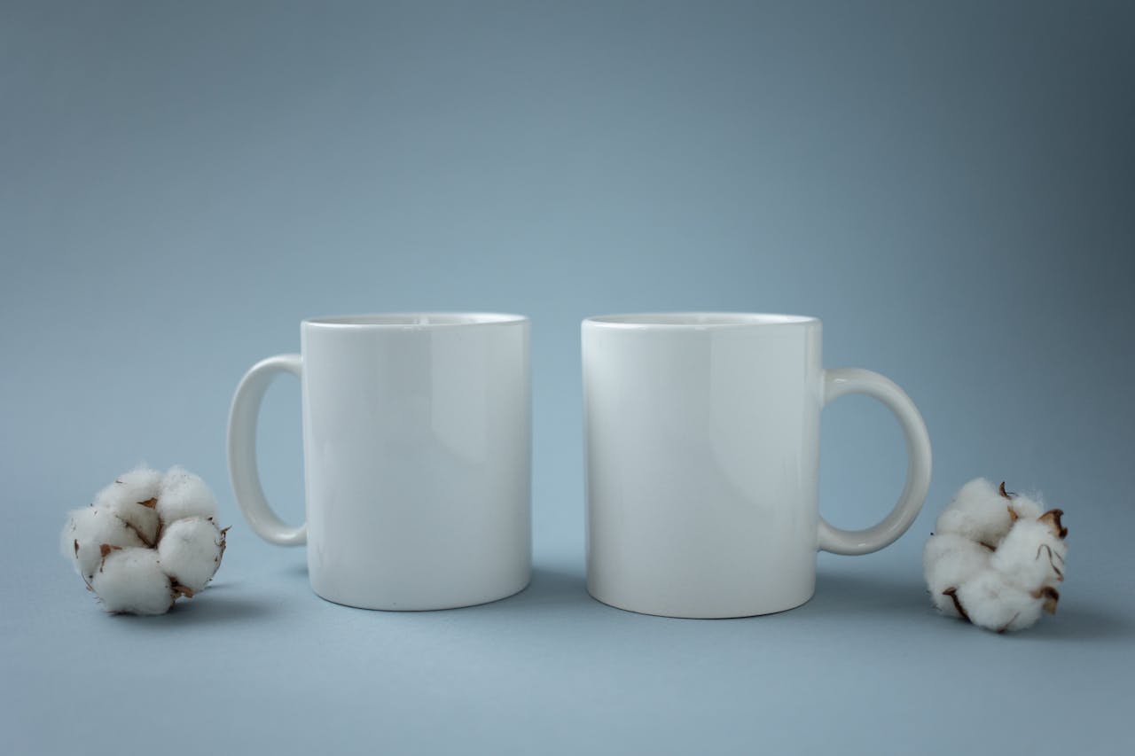 Two white ceramic mugs placed beside cotton flowers on a light blue background, emphasizing minimalism.